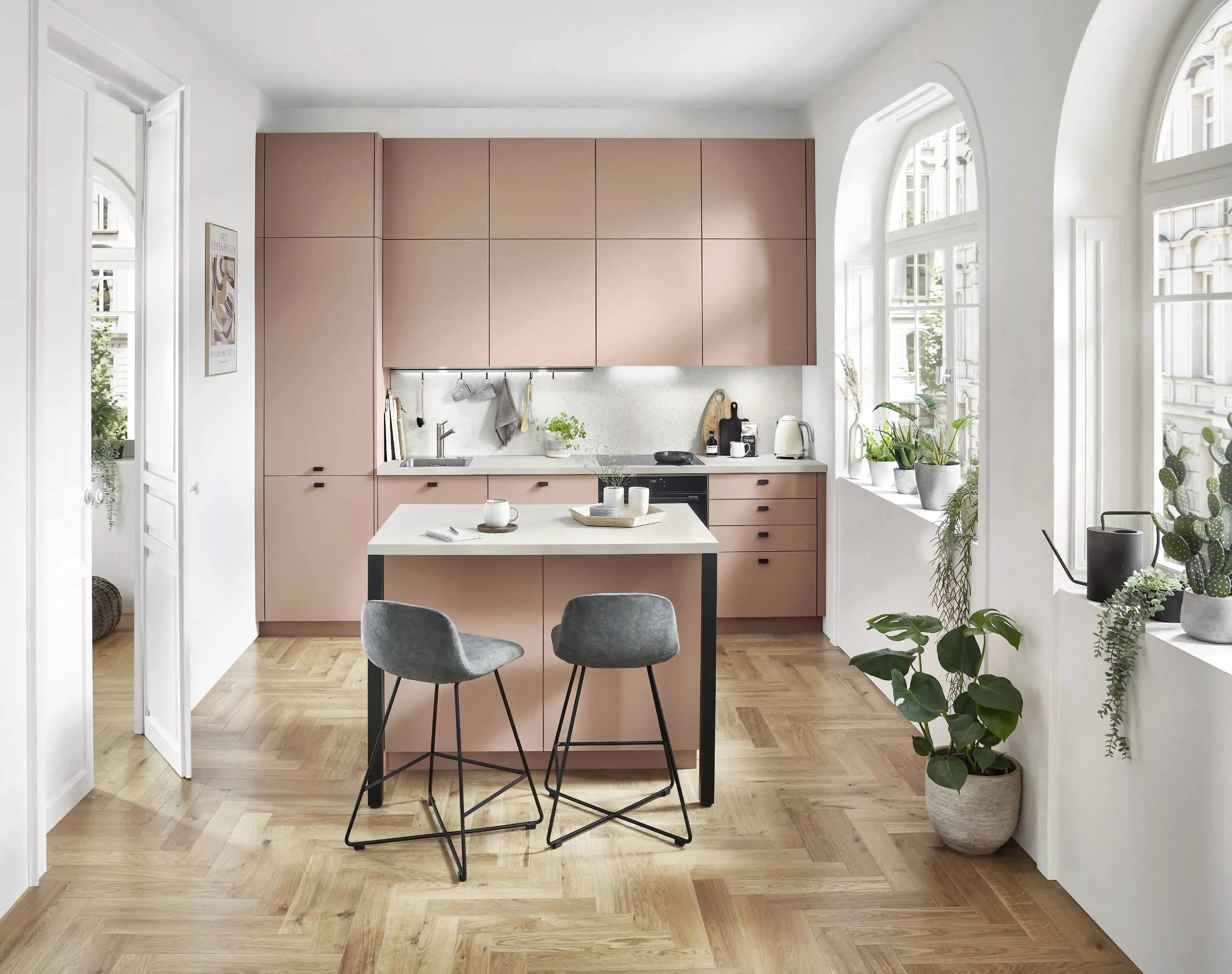 Moderni roosanvärinen designkeittiö, jossa on kahvanomat kaapistot, keittiösaareke, valkoinen työtaso, kalanruotoparketti ja suuret kaari-ikkunat valoisassa tilassa.